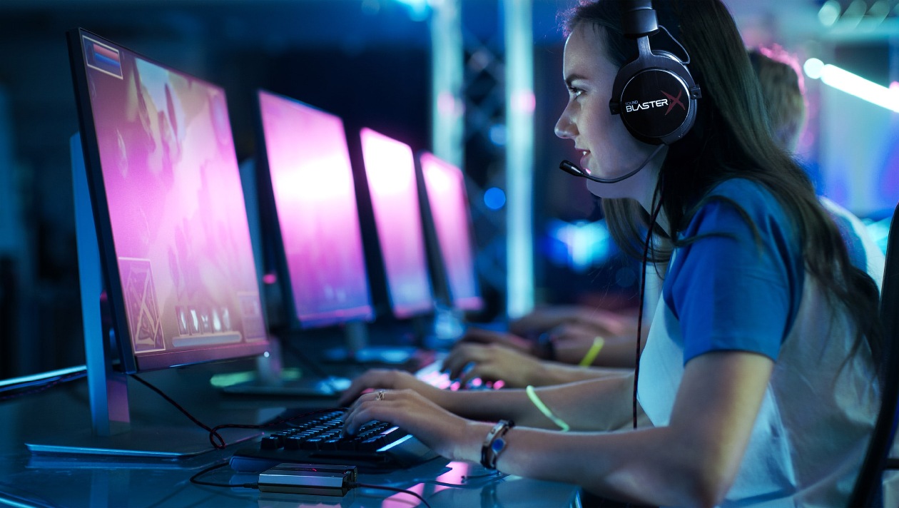 Sound BlasterX G6 vista de perfil sobre mesa em ambiente gamer com iluminação azul e roxa, jogadora usando headset Sound BlasterX