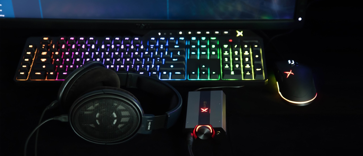 Sound BlasterX G6 vista frontal em mesa escura ao lado de fone de ouvido, com teclado e mouse RGB ao fundo