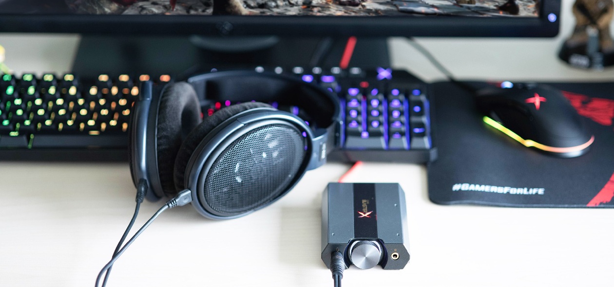 Sound BlasterX G6 vista frontal sobre mesa branca ao lado de fone, teclado e mouse gamer, com mousepad estampado GAMERSFORLIFE
