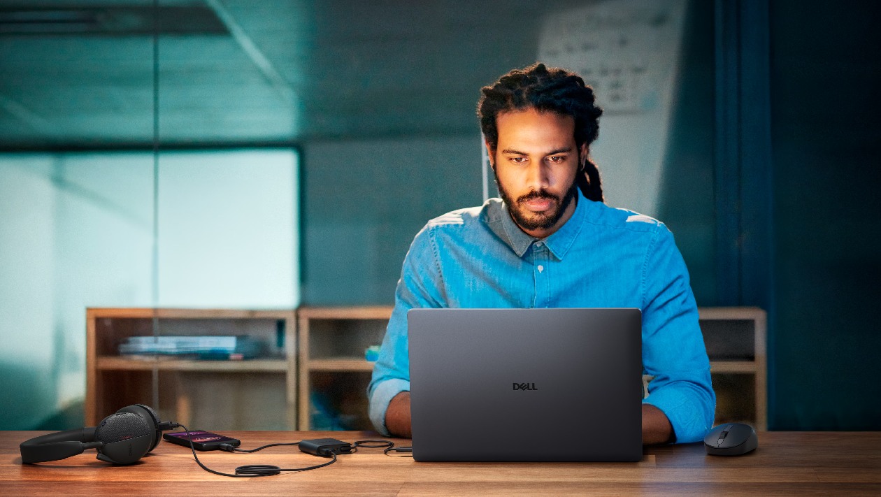Profissional em camisa azul utilizando notebook Dell em mesa de madeira com fones de ouvido, smartphone e mouse conectados ao hub compacto DA225 em ambiente de home office com iluminação suave