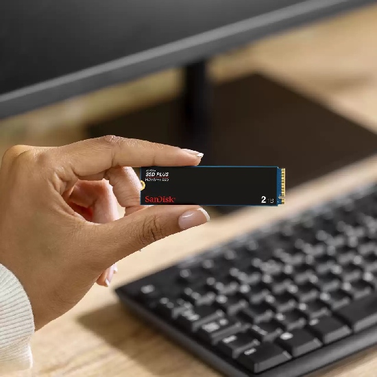 Mão segurando o SSD M.2 NVMe SanDisk SSD PLUS de 2TB em frente a um PC